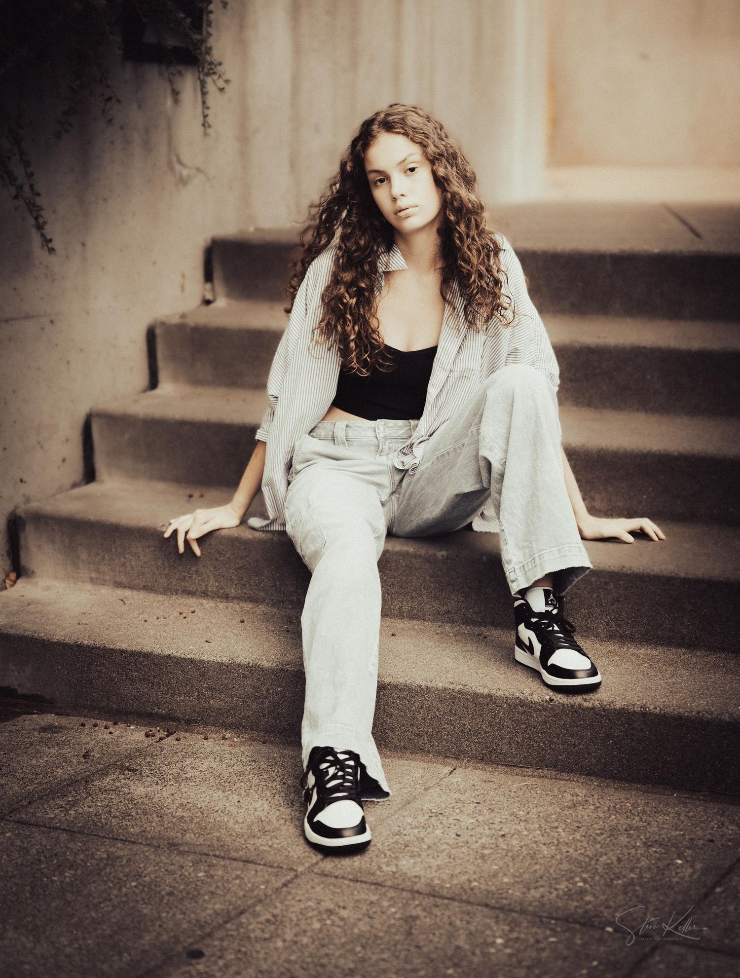 A person in casual attire sitting on concrete steps wearing a light jacket and sneakers in a sepia toned portrait.