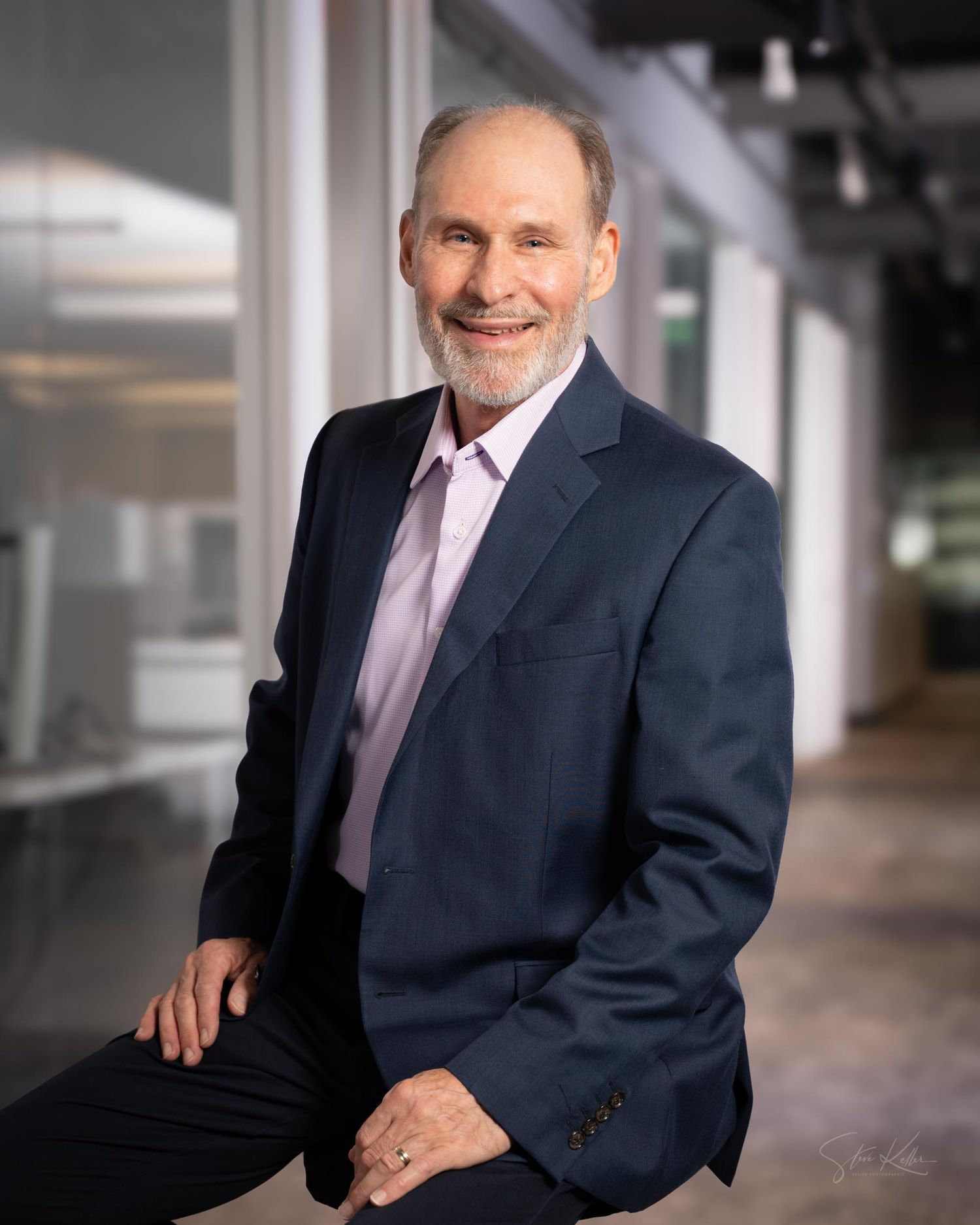 Professional business portrait of someone in a navy suit and pink tie sitting in a modern office setting.