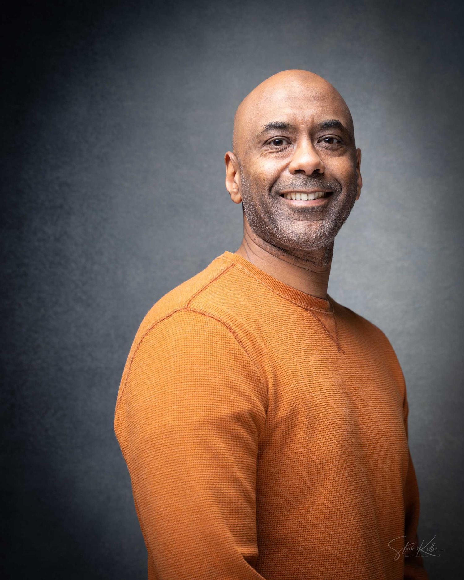 Smiling bald man wearing an orange sweater poses for a professional studio portrait photograph.