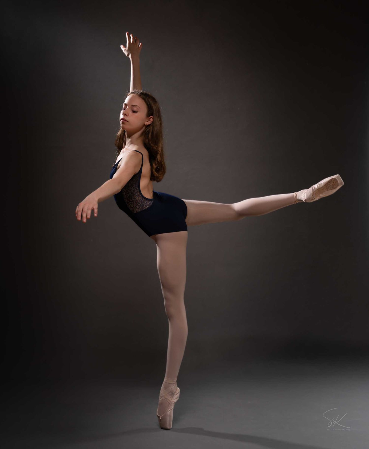 A ballet dancer in a navy leotard performs an arabesque on pointe shoes against a dark studio background.