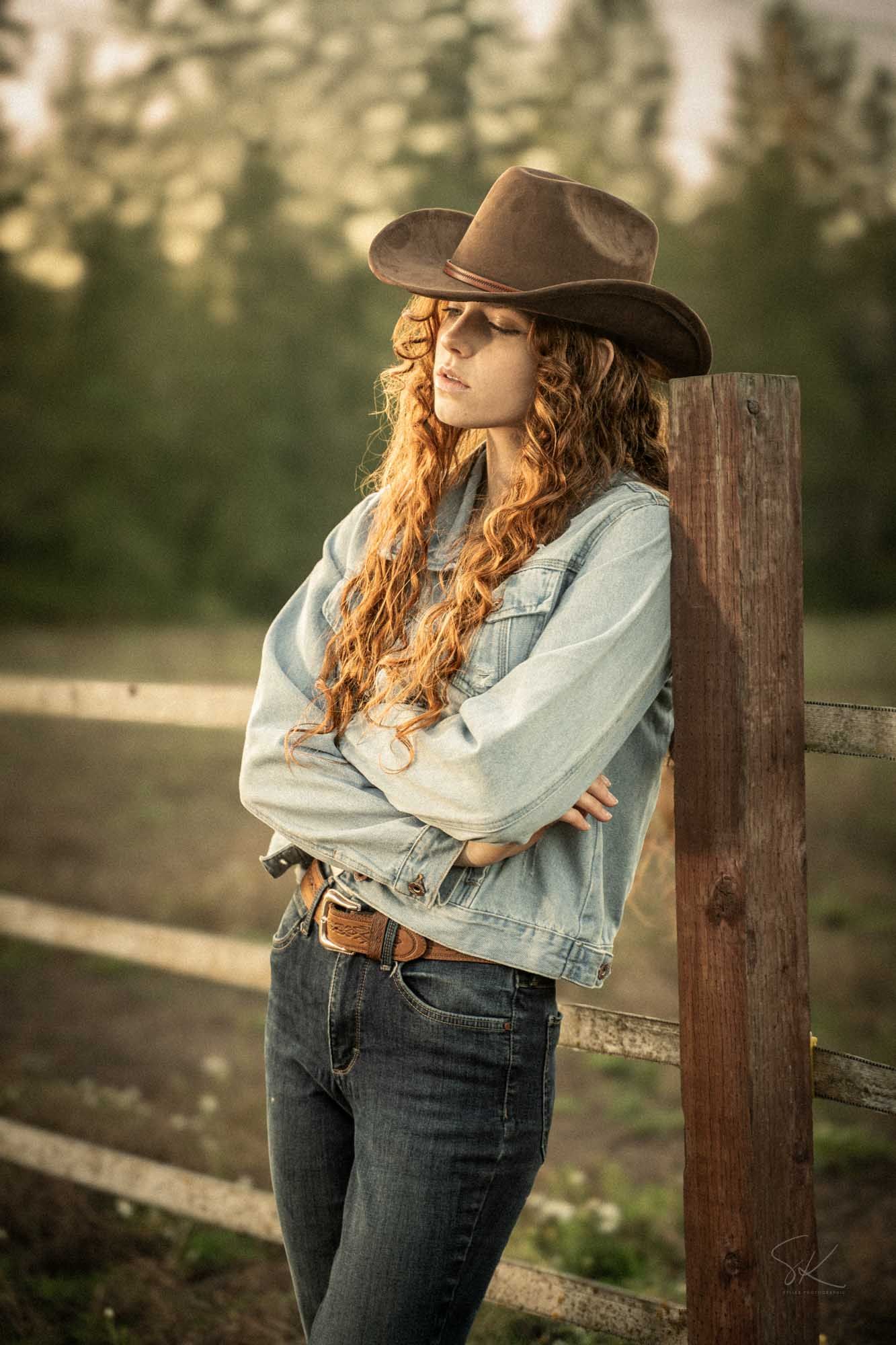 A young woman with curly red hair wearing a cowboy hat, denim jacket, and jeans leans against a wooden fence post.