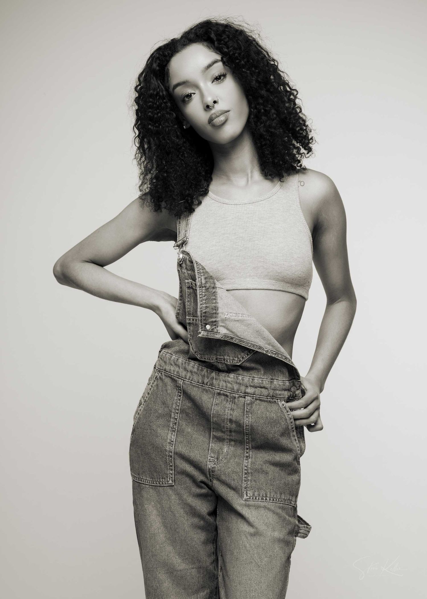 Black and white photo of a young woman with curly hair wearing a crop top and denim overalls pants posing confidently.