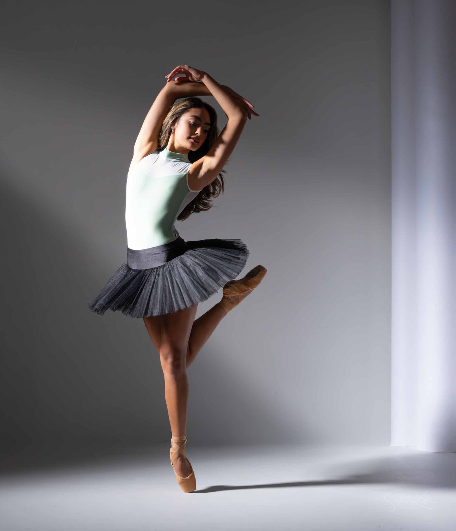 A ballet dancer performs graceful movements in a white tutu against a minimalist gray studio backdrop.