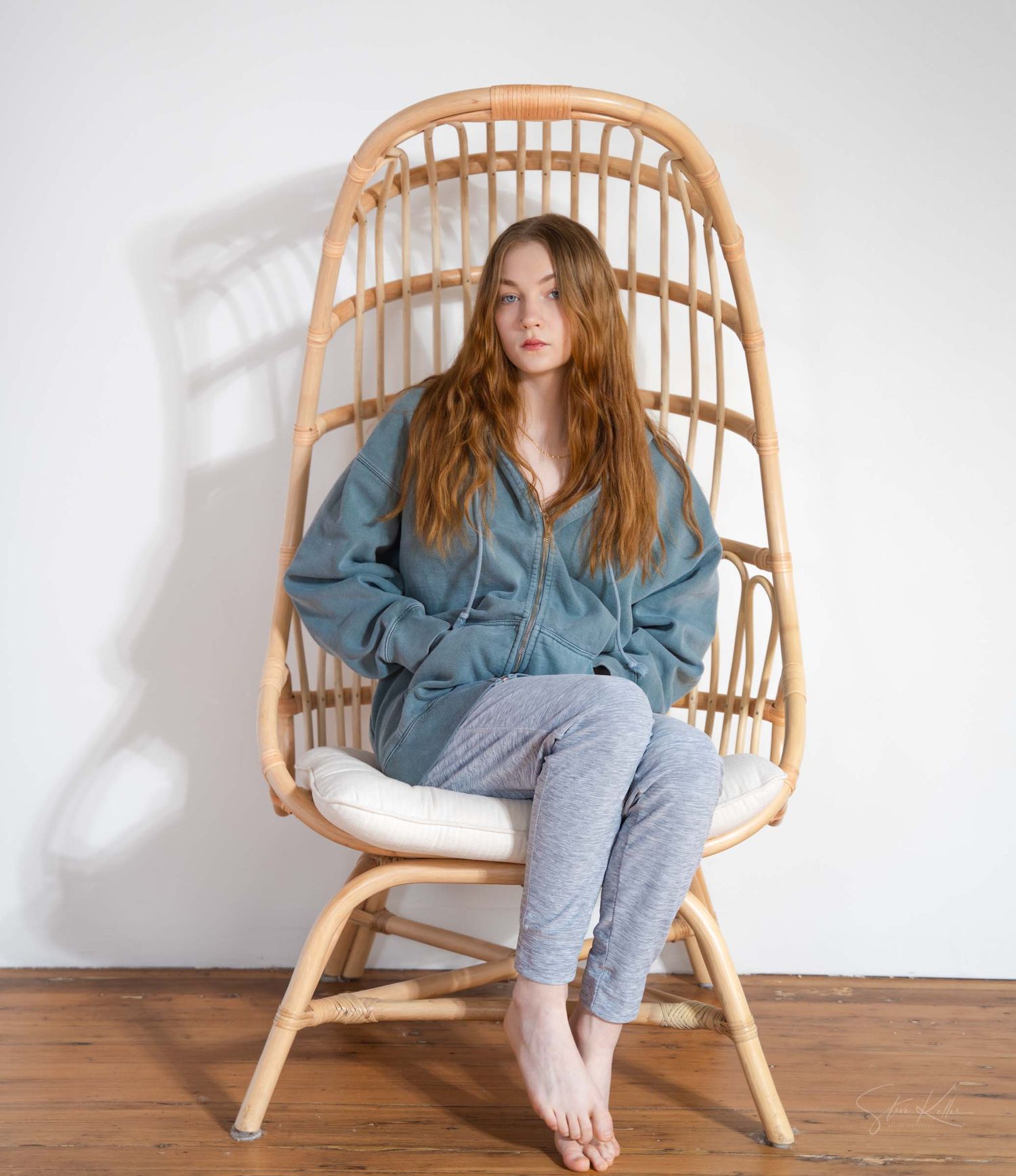 A rattan hanging chair sits against a white wall with someone wearing a denim shirt and gray sweatpants lounging in it.