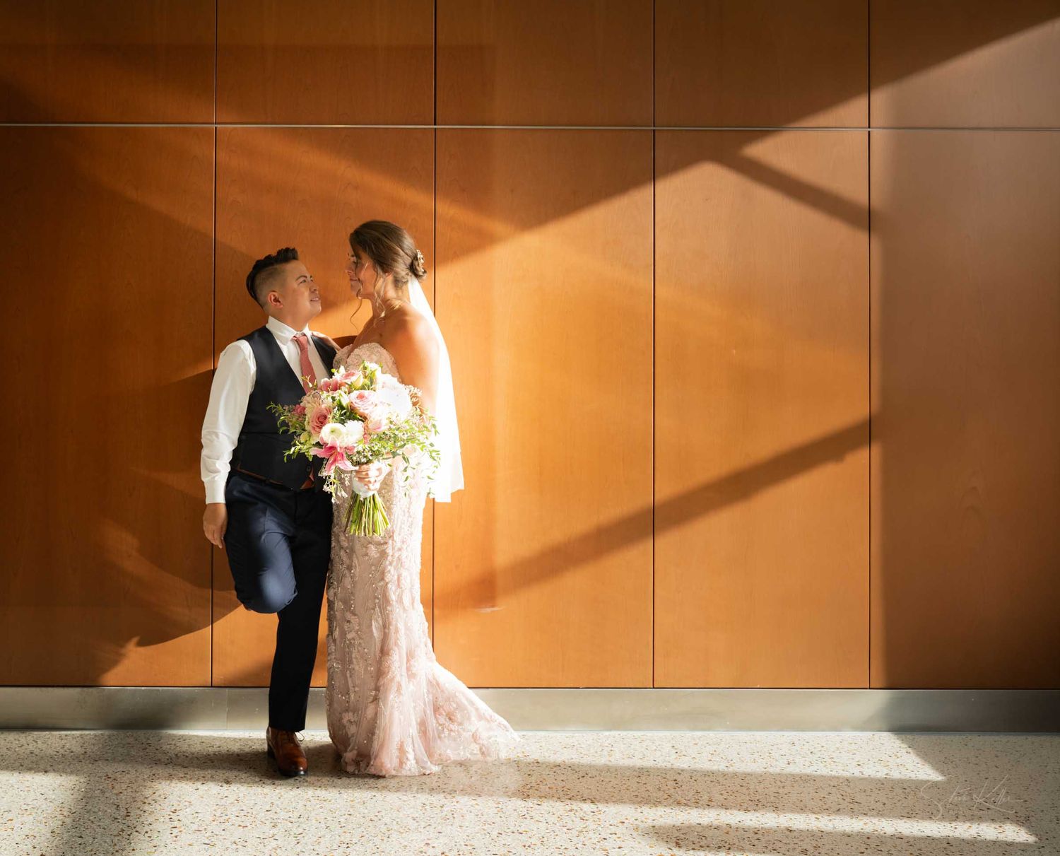 Bride and groom share a romantic kiss in warm golden light against a wood-paneled wall, bride holding a lush floral bouquet.