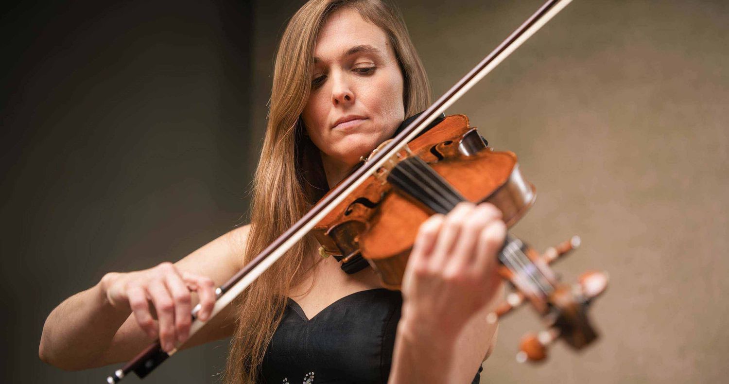 Woman with long hair in a black dress plays a violin with a bow, concentrating during a live classical music performance.