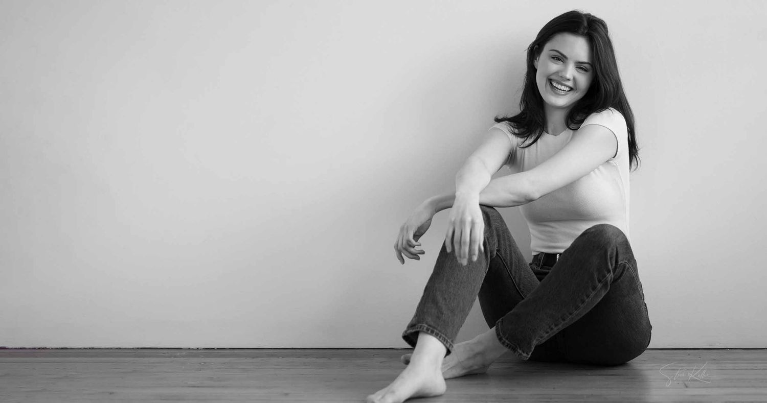 Smiling woman with long dark hair sits barefoot on a wooden floor against a plain wall in a black and white portrait.