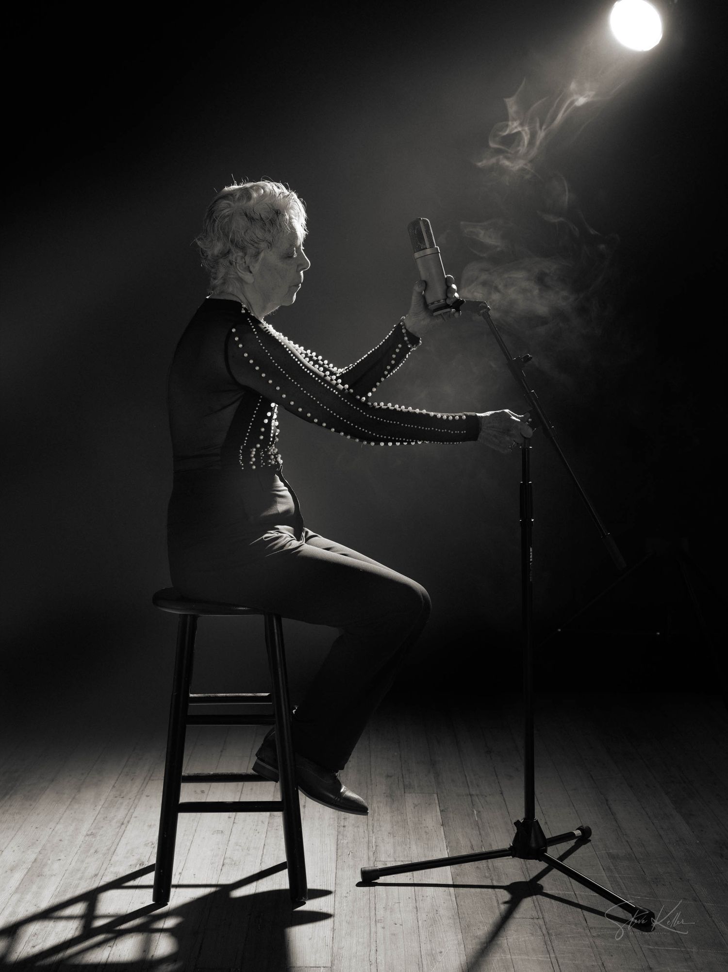 Dramatic black and white side view of a performer on a stool under a spotlight with smoke effects on a dark stage.