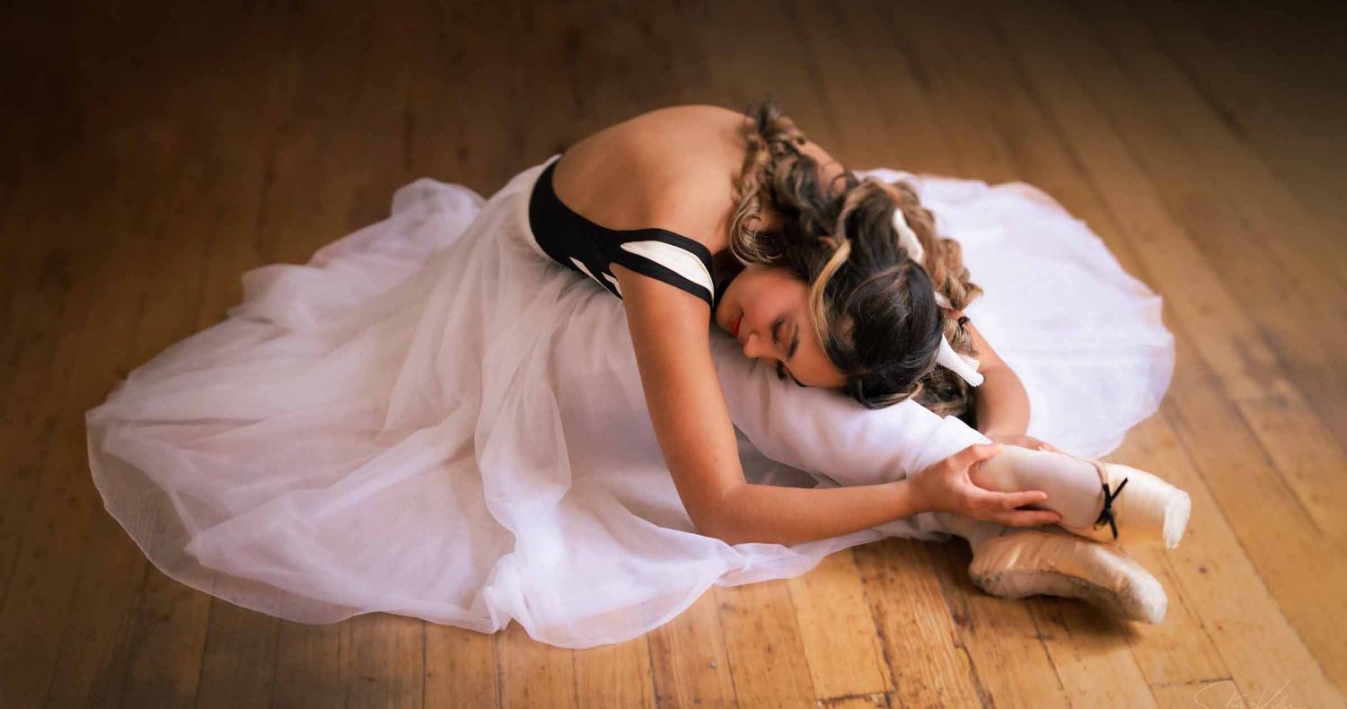Ballerina in a flowing white tutu sits on a wooden studio floor, stretching forward over her legs while adjusting her pointe shoe.