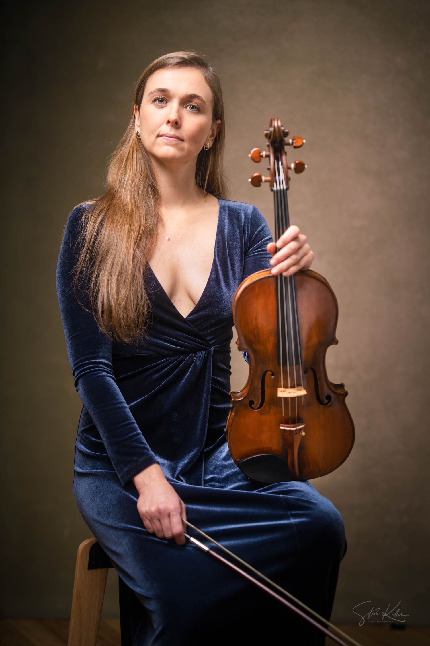 A musician in a navy velvet dress poses with a violin against a dark backdrop in a professional portrait series.