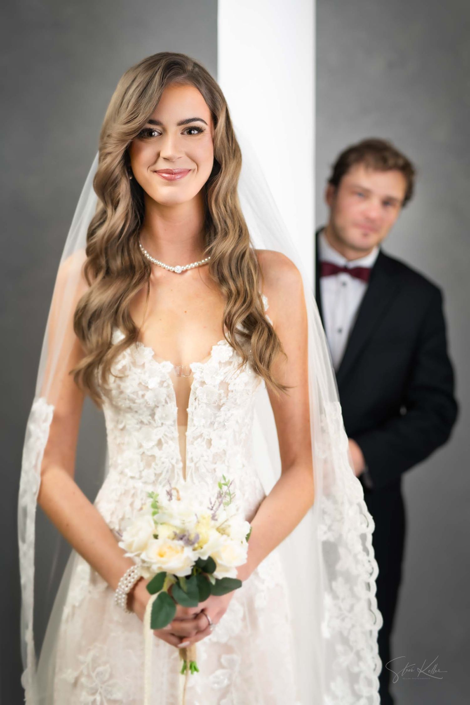 A bride in a white lace wedding dress with plunging neckline holds a white floral bouquet in a studio setting.