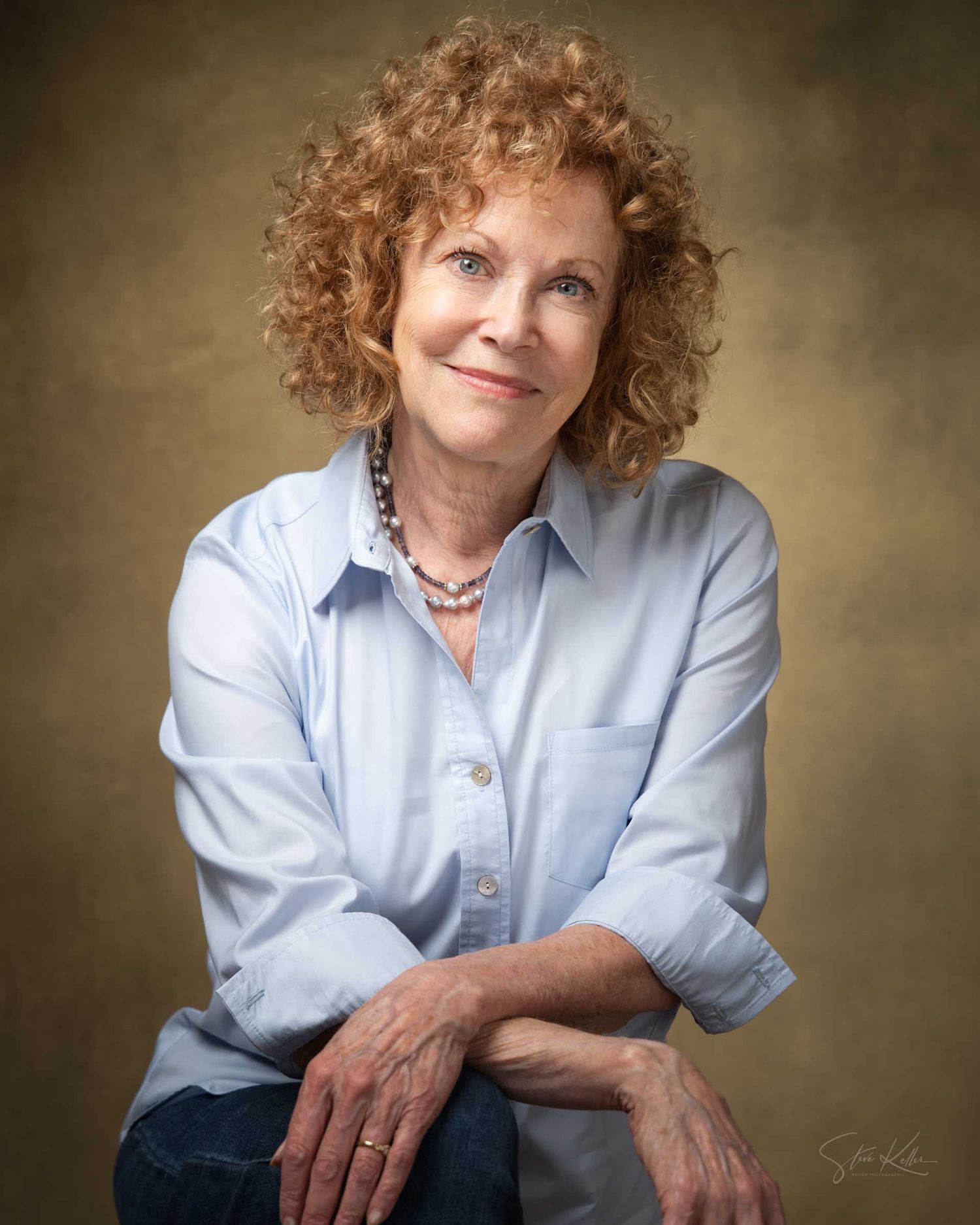 A smiling woman with curly red hair wearing a light blue shirt and pearl necklace poses for a professional portrait.