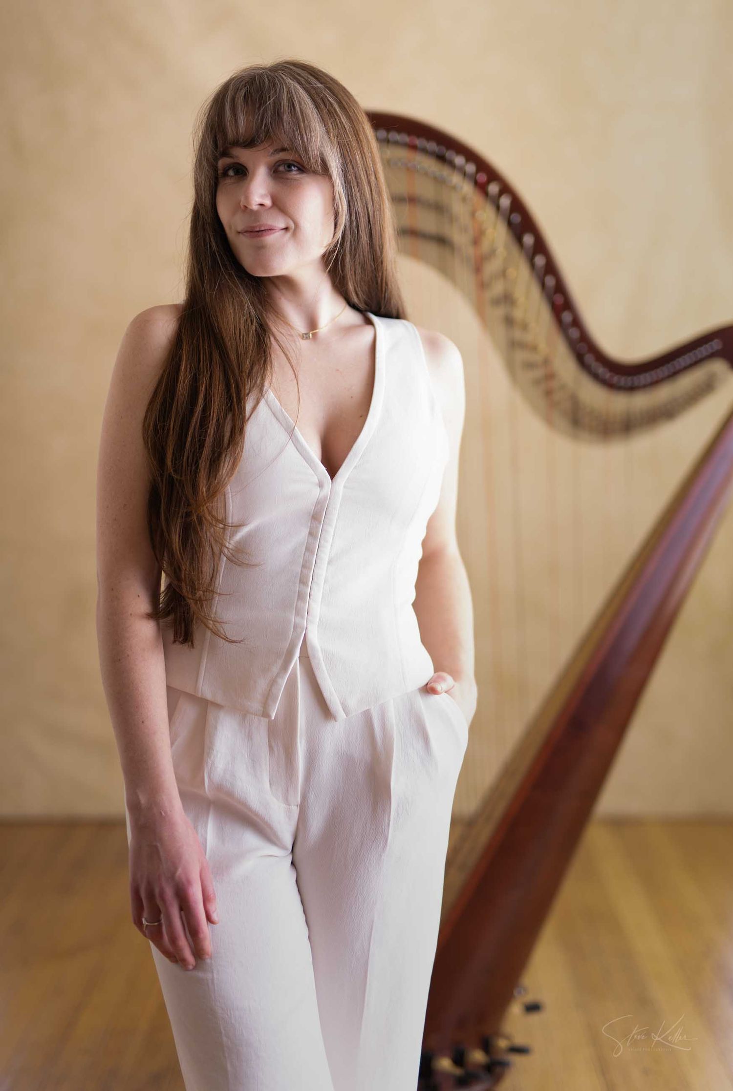 Woman in white jumpsuit posing beside a wooden harp on a warm-toned wooden floor background.