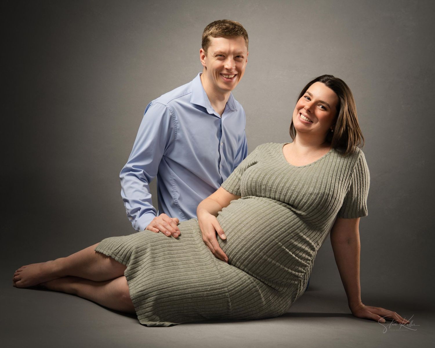 A couple poses together during a maternity photo shoot on a gray backdrop, with the woman wearing a ribbed gray dress.