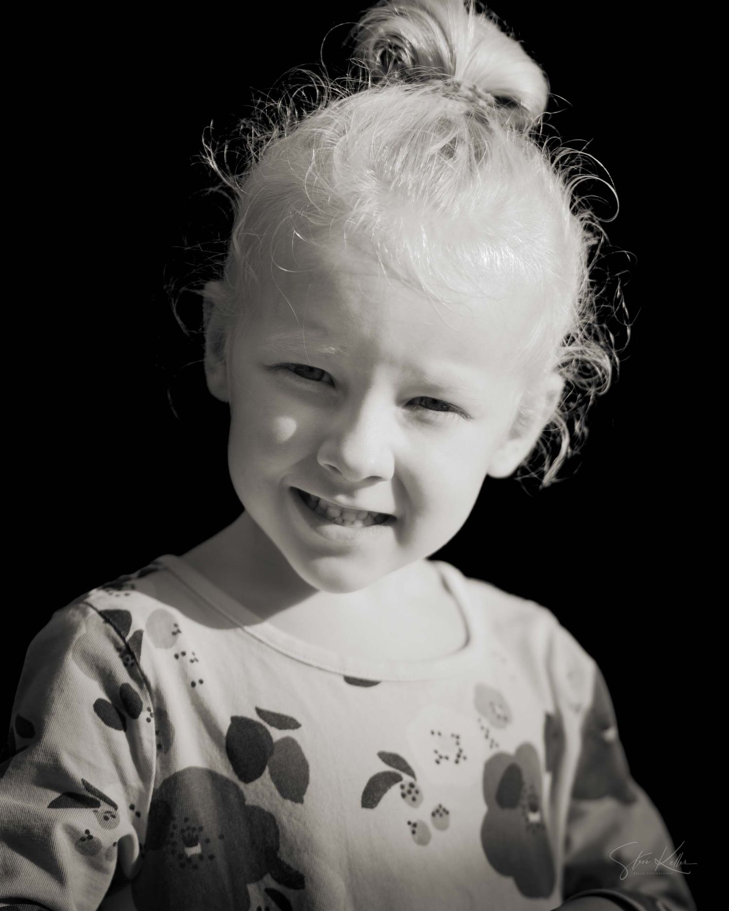 Black and white portrait of a smiling toddler with wispy hair against a dark background.
