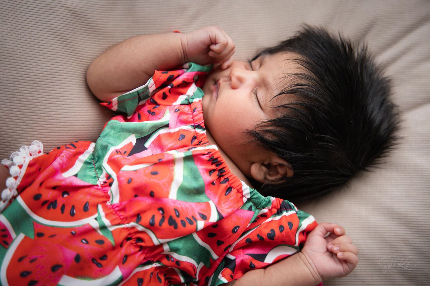 Baby sleeping peacefully on beige bedding while wearing a vibrant red watermelon print outfit.