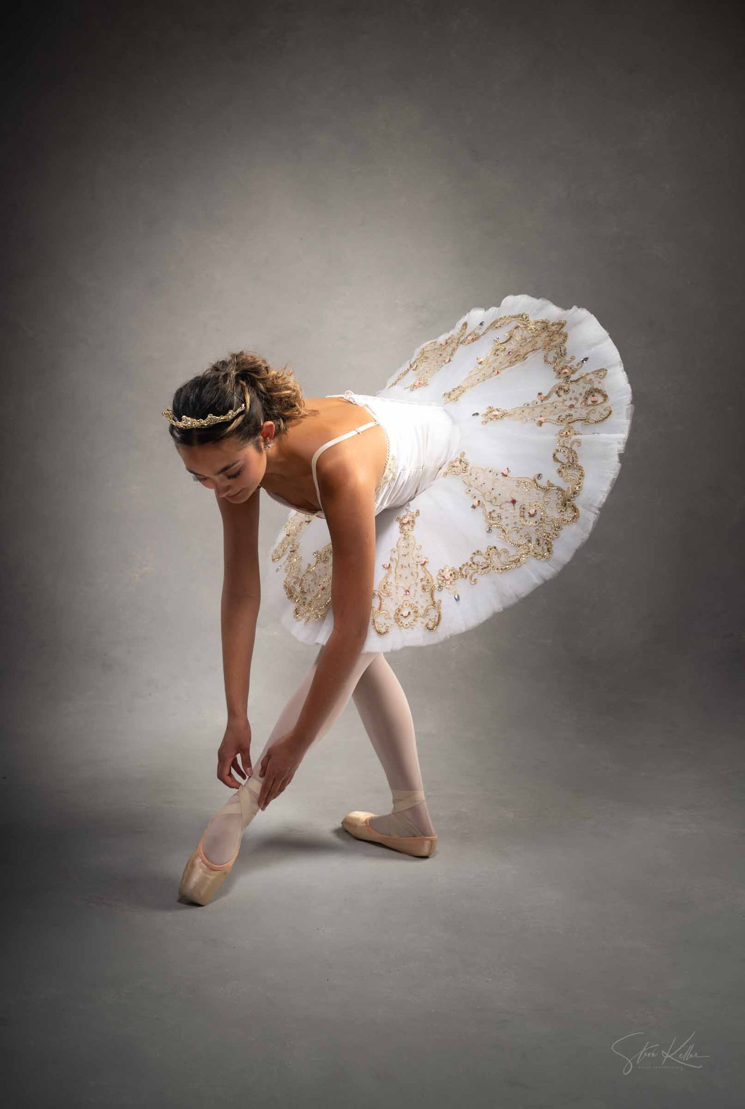 A graceful dancer in a white tutu performs a delicate bending movement on a dark studio floor.