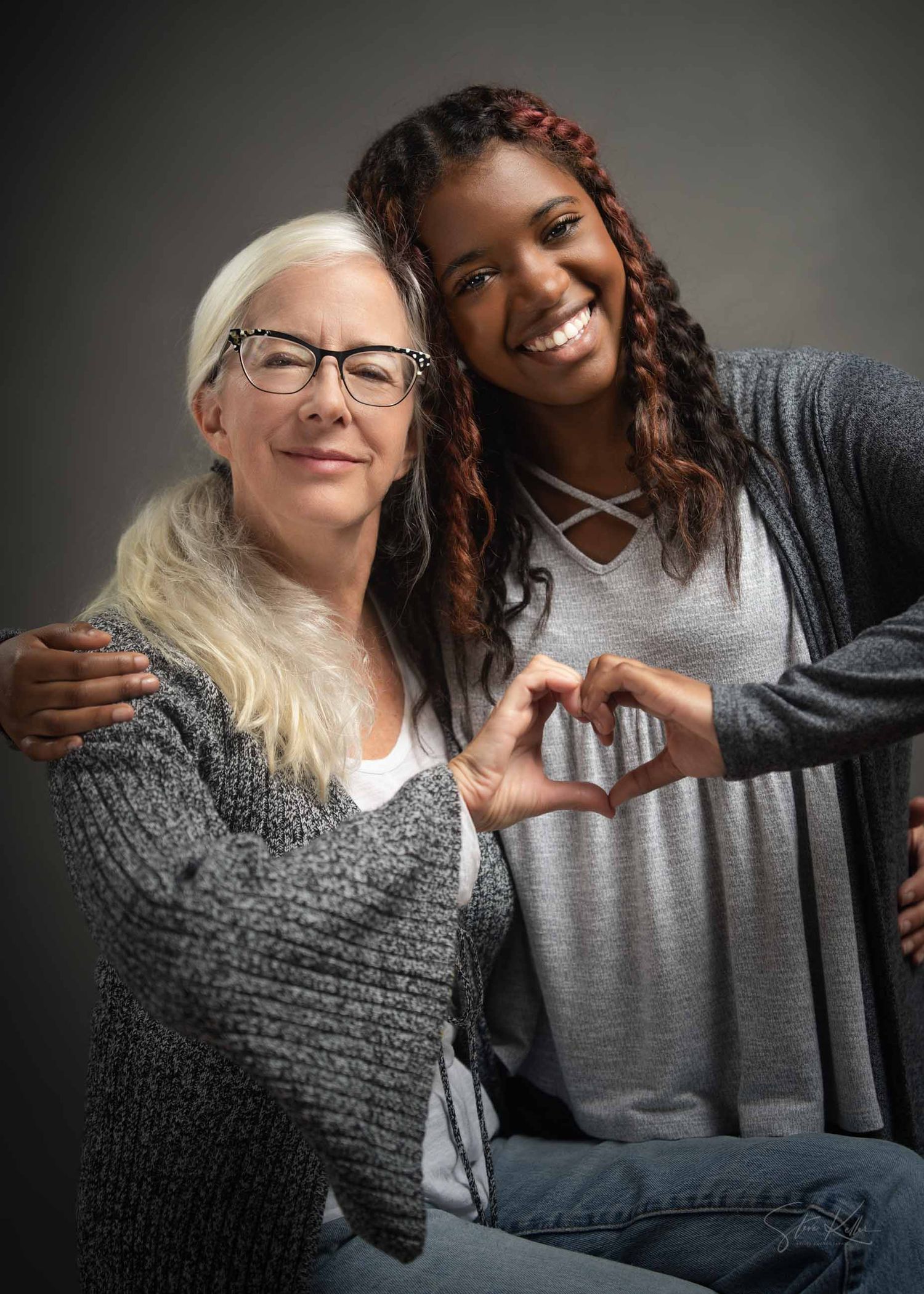 Two people in gray sweaters share a joyful moment while making a heart shape with their hands.