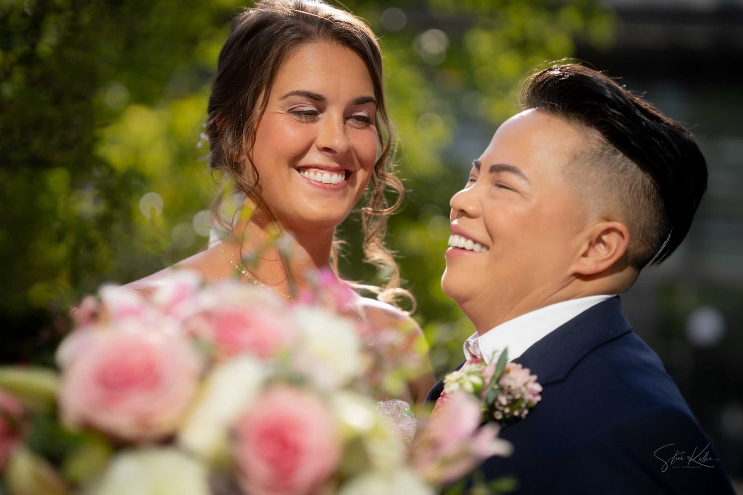 Two brides share a joyful laugh outdoors, one in a pink gown and the other in a navy suit, with a floral bouquet in the foreground.