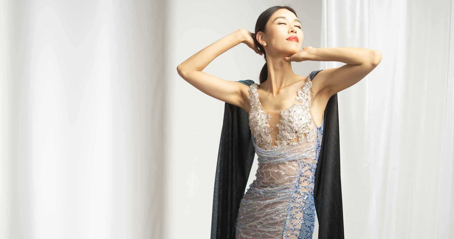 Elegant woman poses with arms raised in a sparkling sheer embellished evening gown with a black cape against a soft white backdrop.