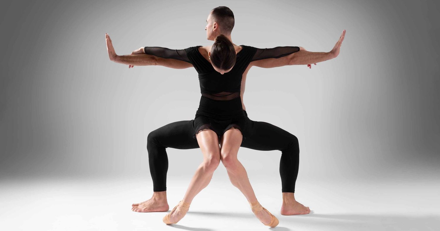 Two dancers in black outfits perform a symmetrical pose, their bodies combined to appear as one figure with outstretched arms and four legs.