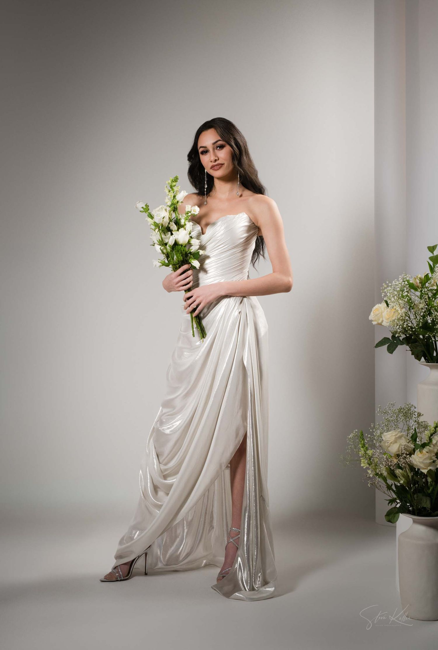 Ethereal maternity photoshoot featuring flowing white fabric and delicate floral arrangements in a minimalist studio setting.