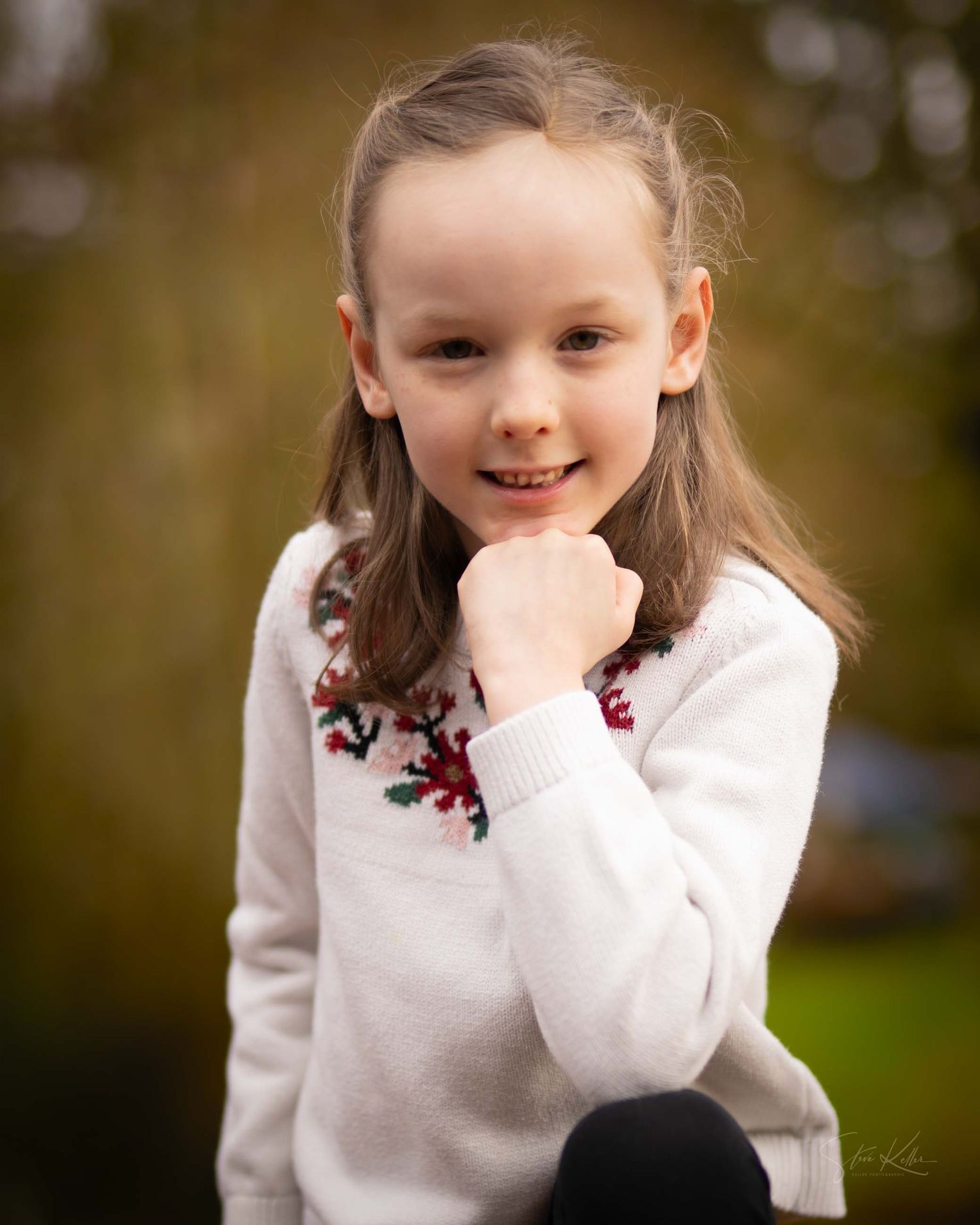A young child in a white sweater with red accents smiles playfully in an outdoor setting with a blurred background.