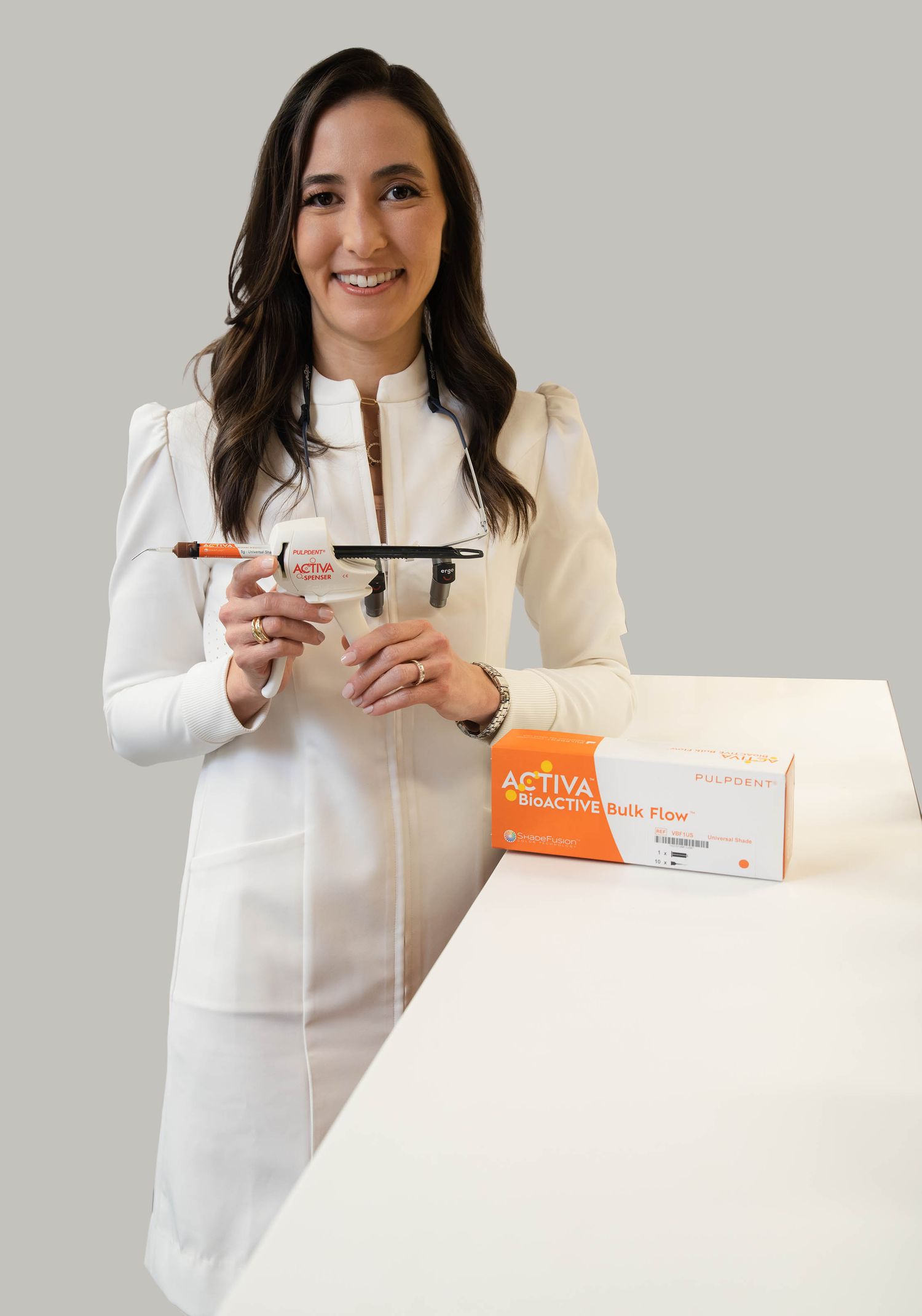Dental professional in white coat holding ACTIVA BioACTIVE Bulk Flow syringe beside product box on white surface.