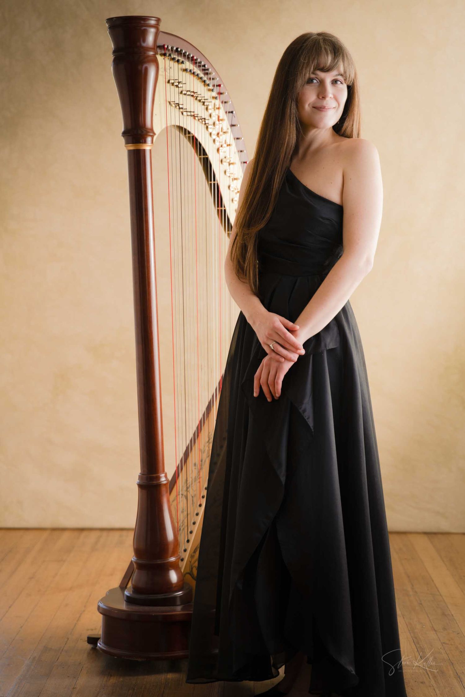 A woman in a black strapless gown stands elegantly beside a tall wooden concert harp.