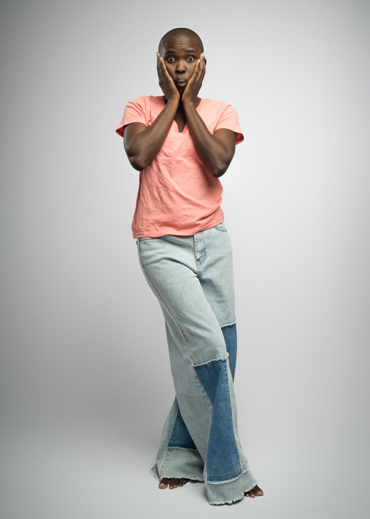 Person in a peach-colored t-shirt and patchwork jeans expressing shock with hands on their cheeks against a gray background.