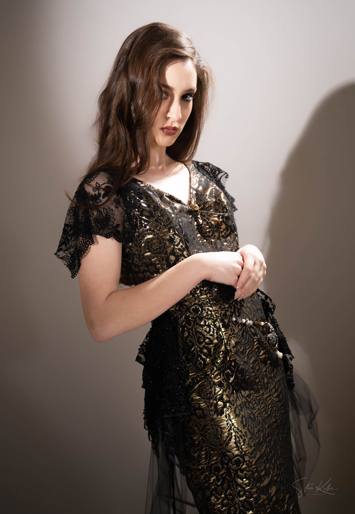 Model wearing ornate black and gold lace dress poses gracefully in studio lighting.