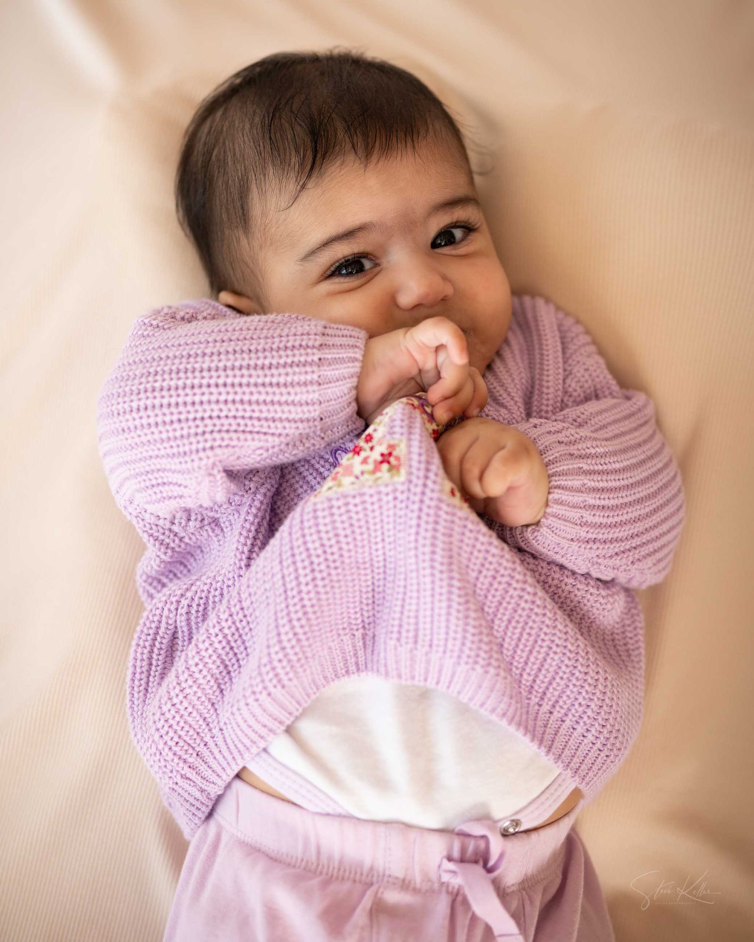 Adorable baby in a lavender knit sweater and pink pants, smiling while chewing on fabric, lying on a beige background.