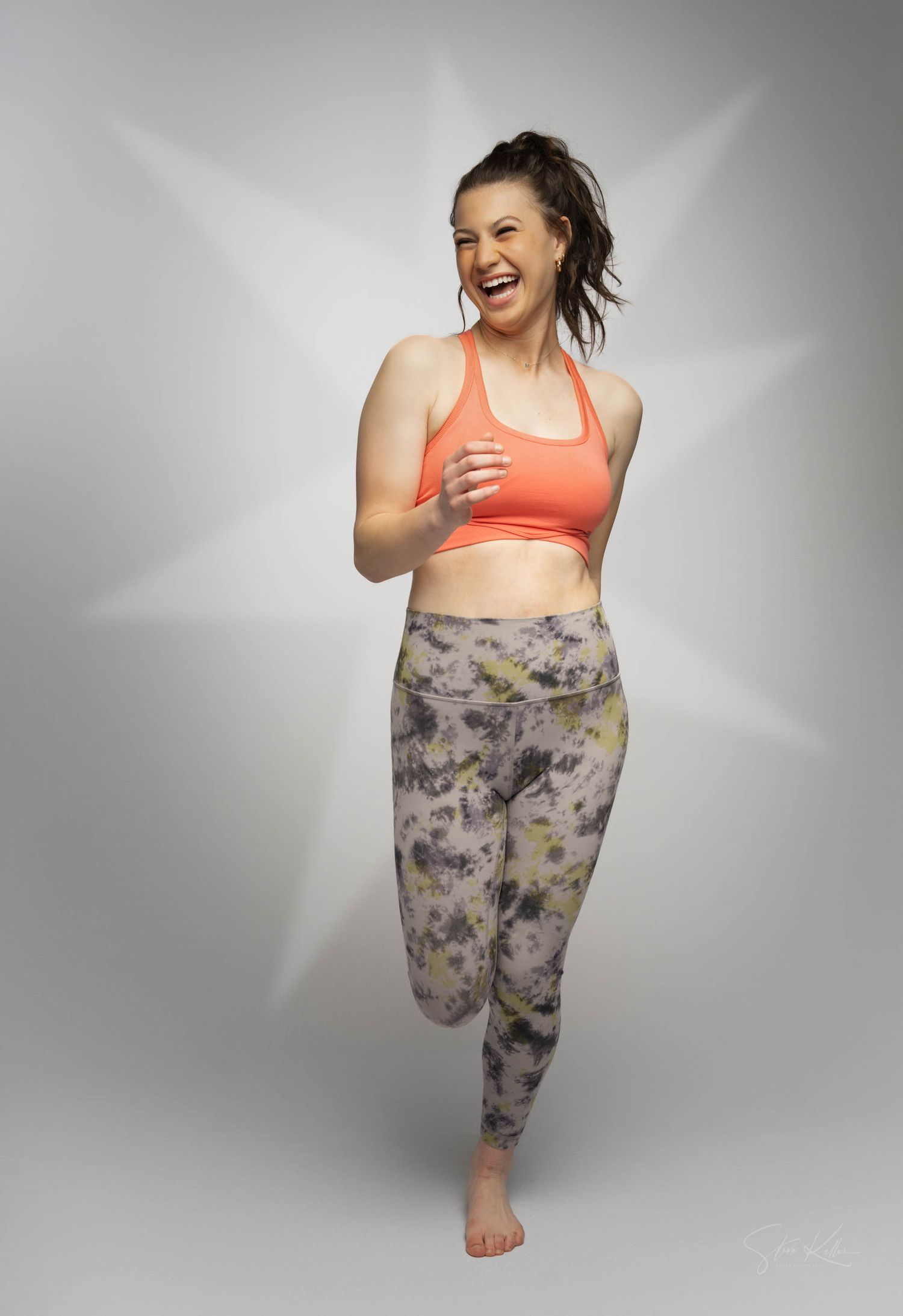 Athletic fashion model in coral sports bra and tie-dye leggings laughing joyfully while striking a dynamic pose against gray background.