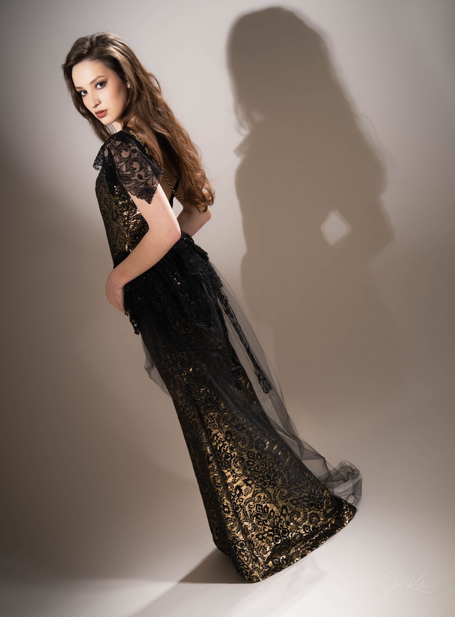 Model in elegant black and gold lace evening gown poses dramatically with shadow cast on beige wall.