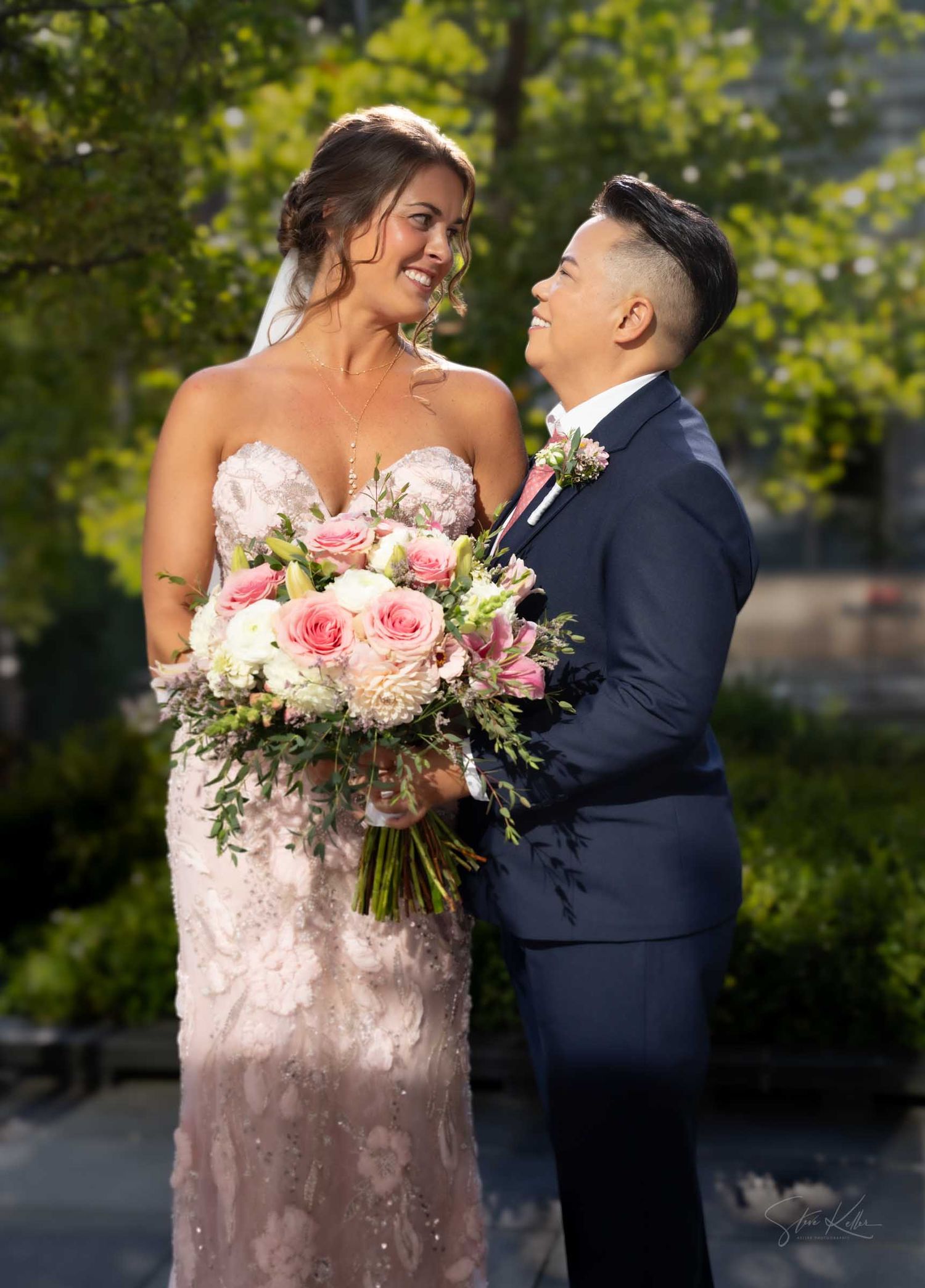 Couple in wedding attire smiling at each other outdoors, bride holding pink and white rose bouquet, groom in navy suit.