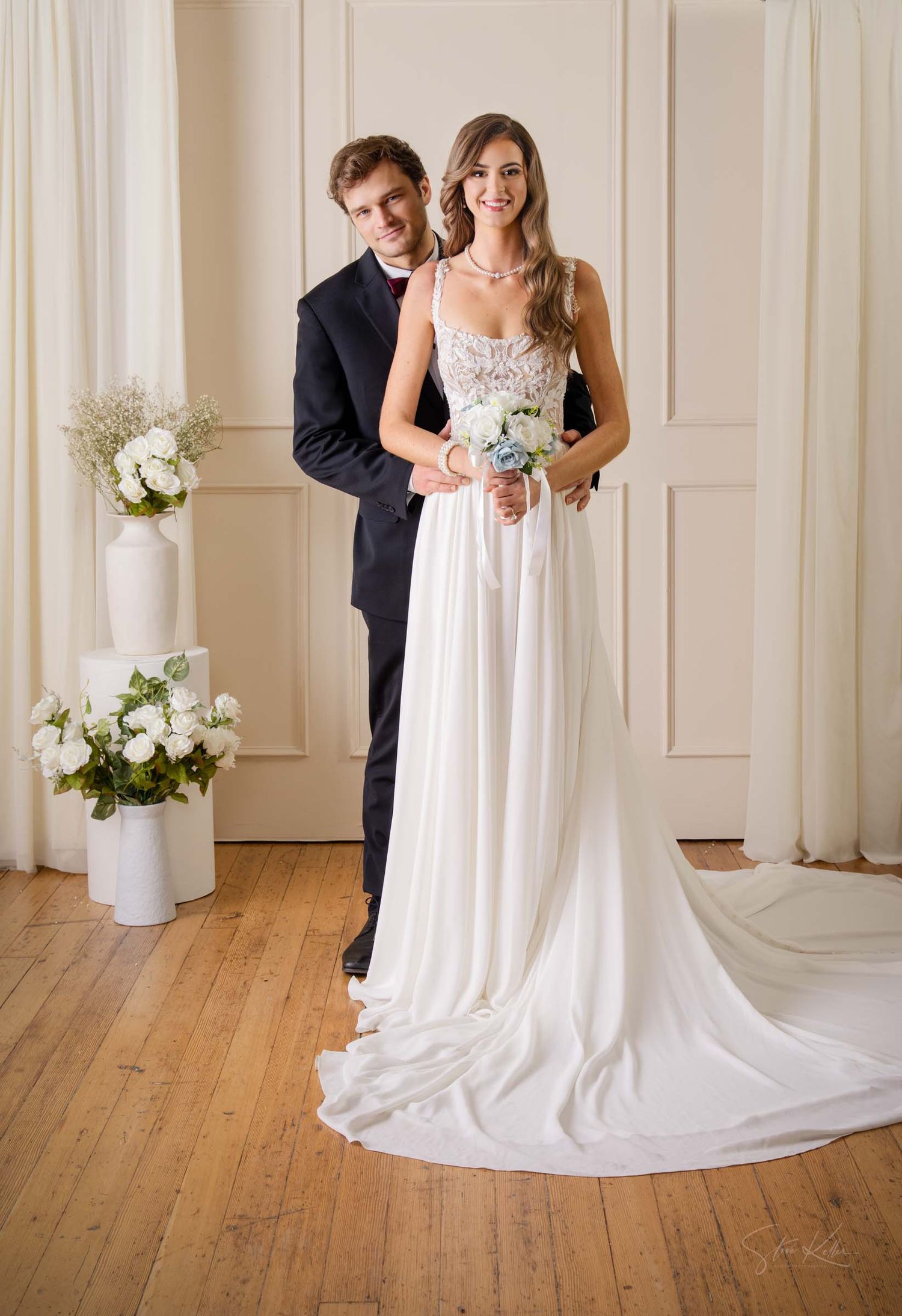 Elegant couple poses in formal wedding attire with white floral arrangements in a classic indoor setting.