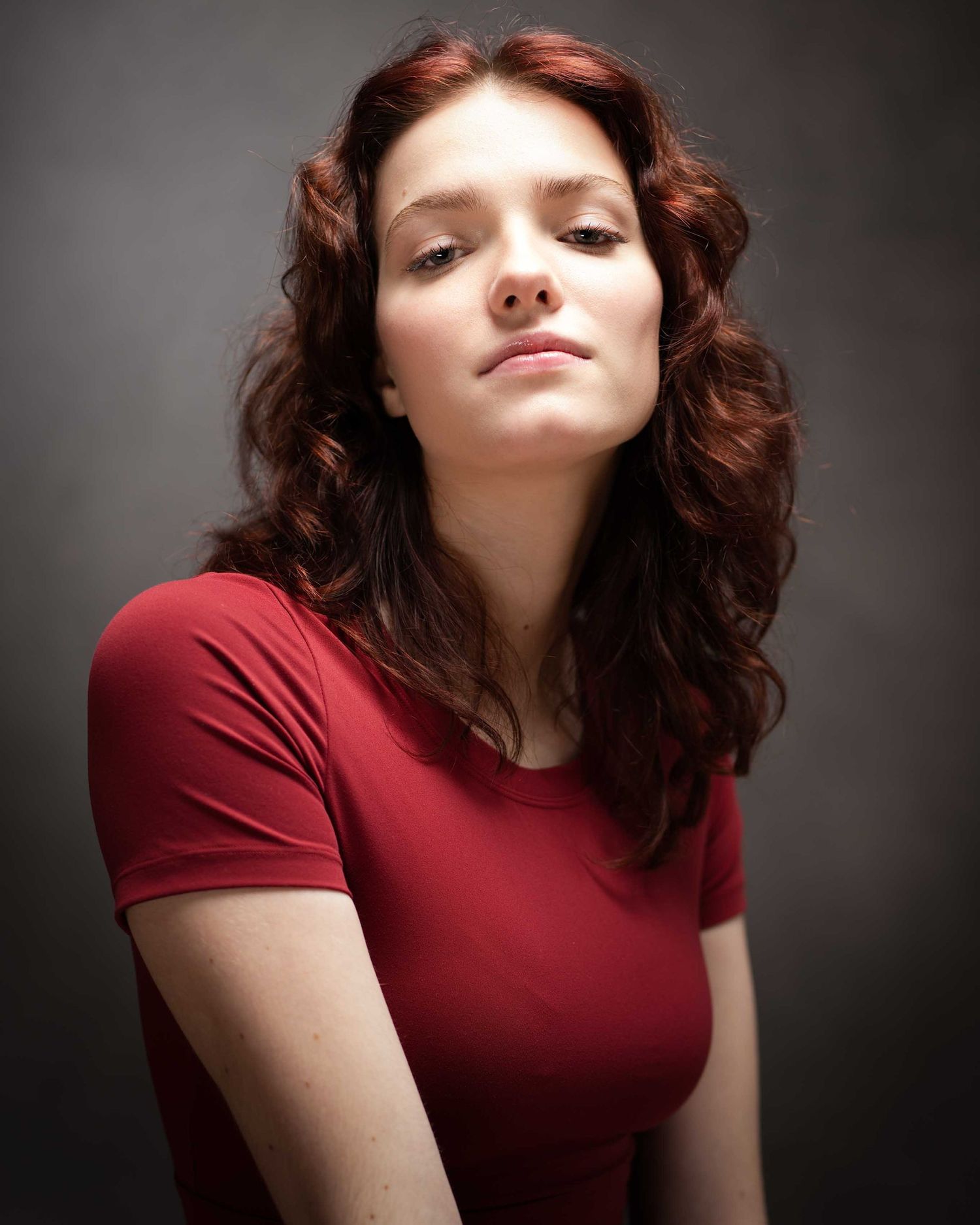 Portrait of a person with wavy auburn hair wearing a red t-shirt against a dark background with dramatic lighting.