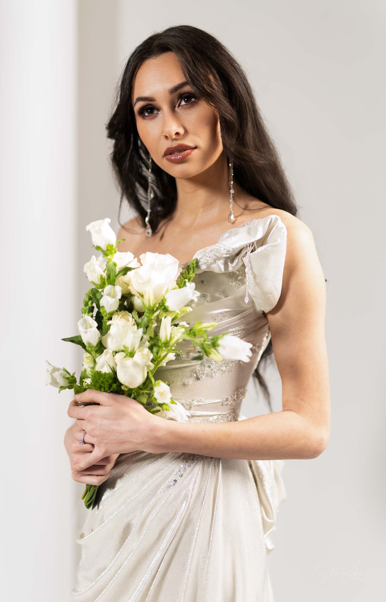 A bride in an elegant ivory gown holds a beautiful white rose bouquet while posing against a light background.