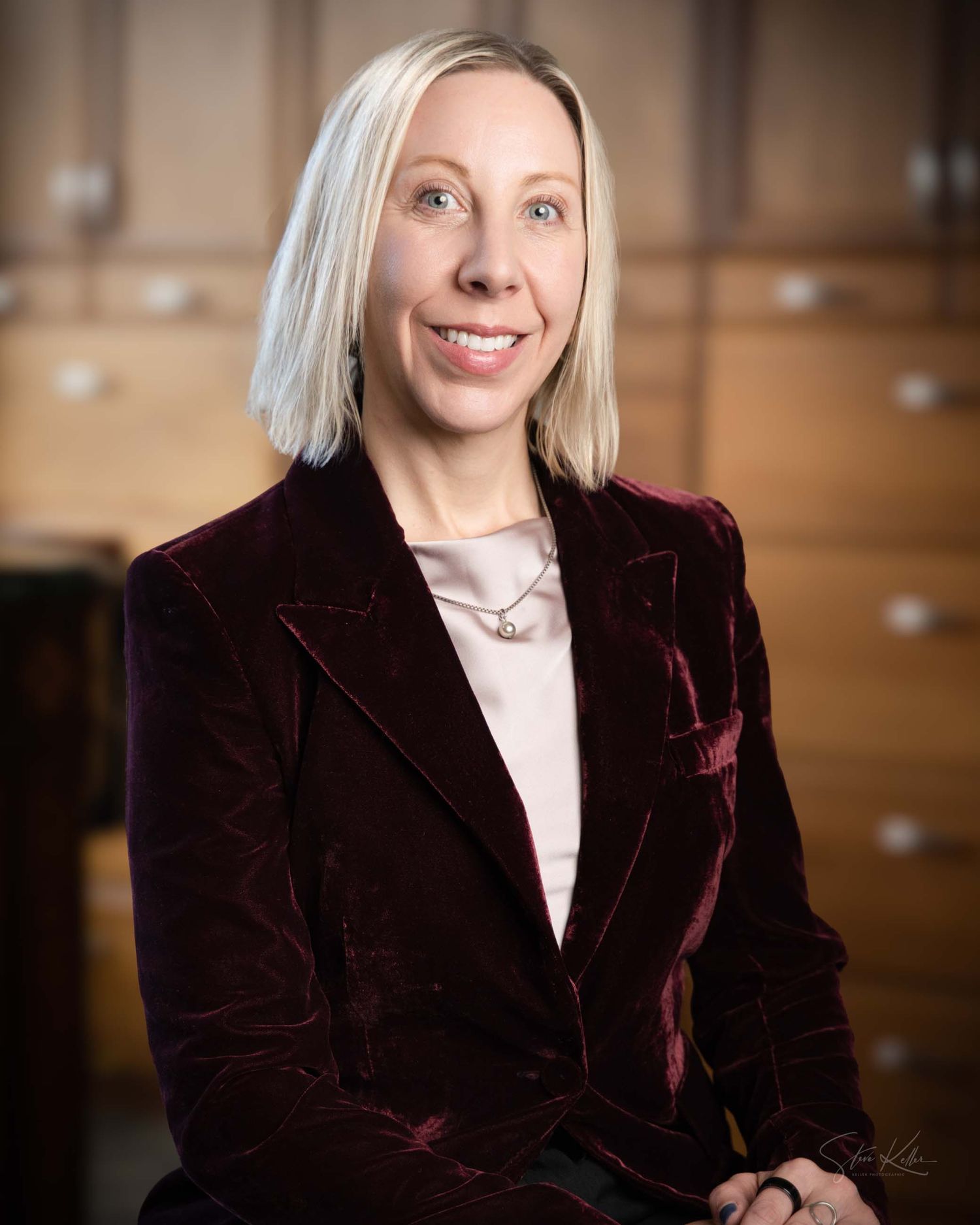 Professional portrait in burgundy velvet blazer and white top against warm brown background with soft lighting.