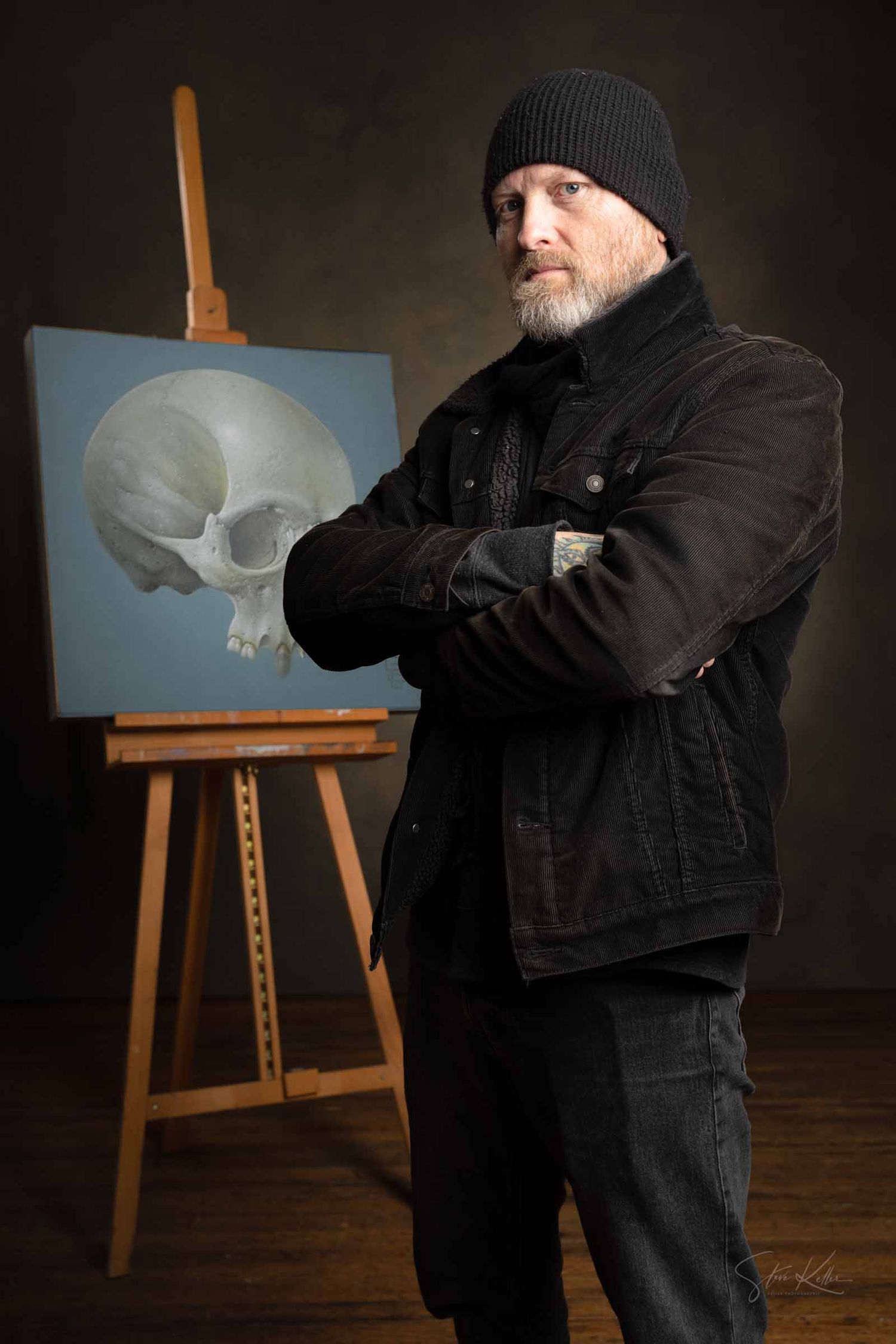 Artist in black clothing and beanie stands with arms crossed next to an easel displaying a skull painting in a dark studio.