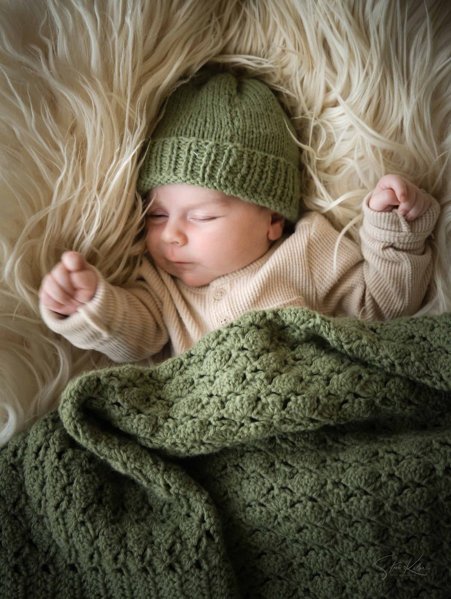 Sleeping baby wrapped in a cozy green knit blanket with matching hat against white fur background.