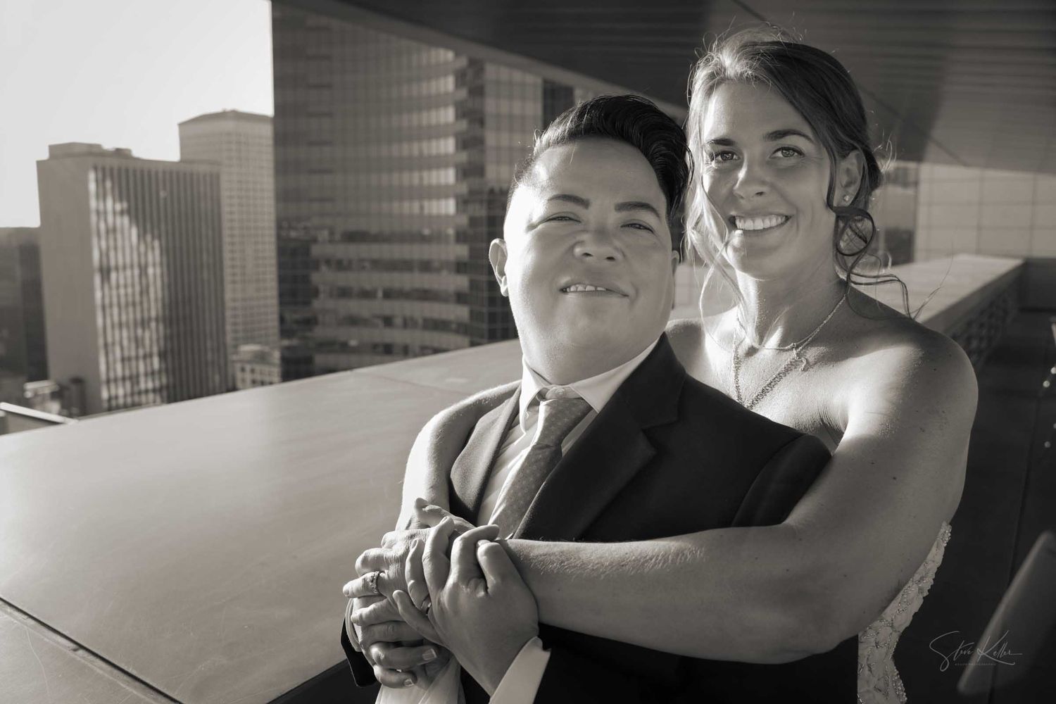 Black and white wedding photo of a couple embracing on a rooftop with city buildings in the background.
