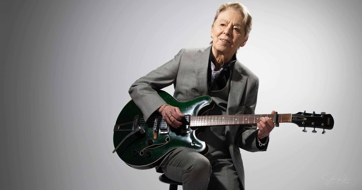 Older man in a gray suit sits on a stool playing a green hollow-body electric guitar against a plain light gray studio backdrop.