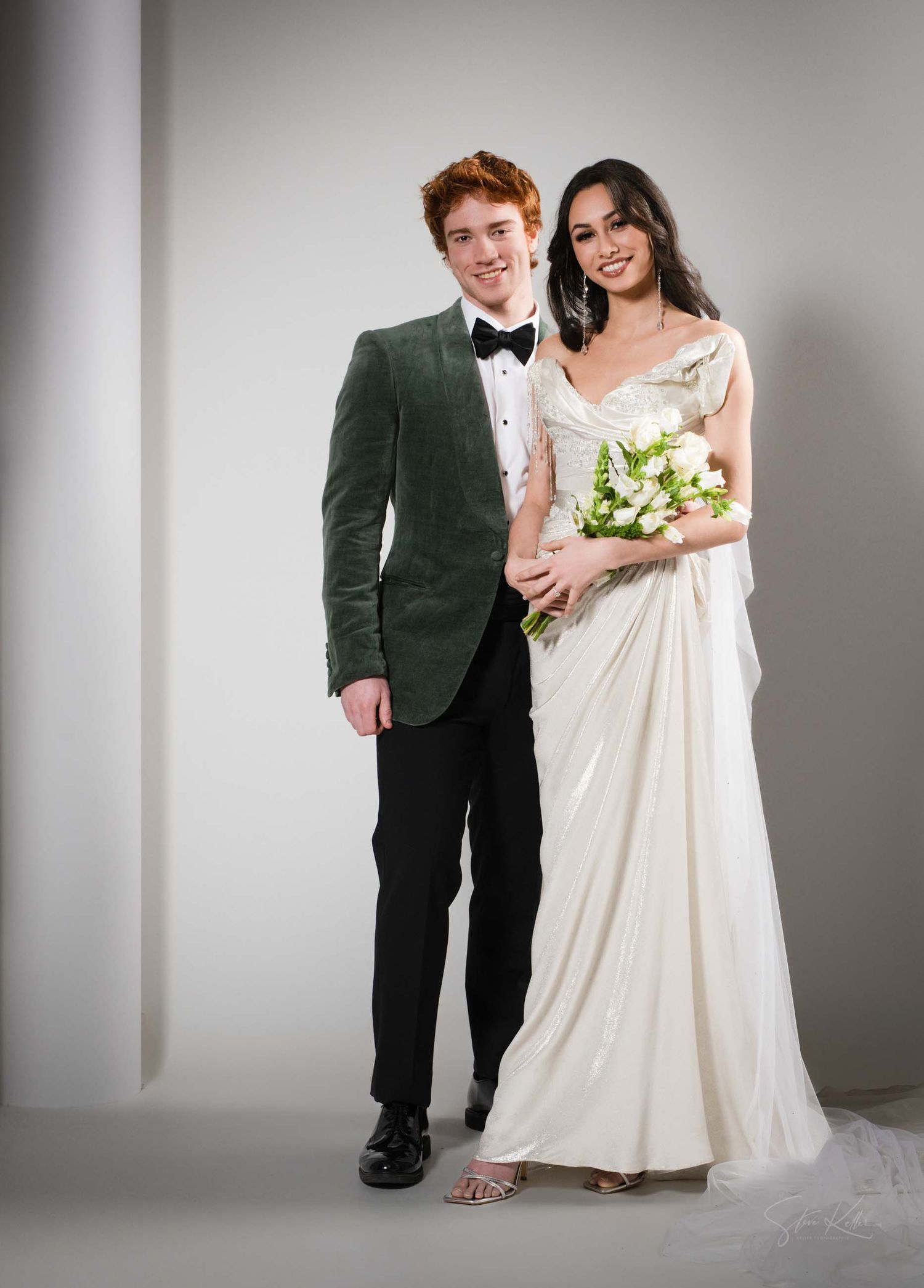 A couple in wedding attire poses together against a light gray backdrop in a studio setting.