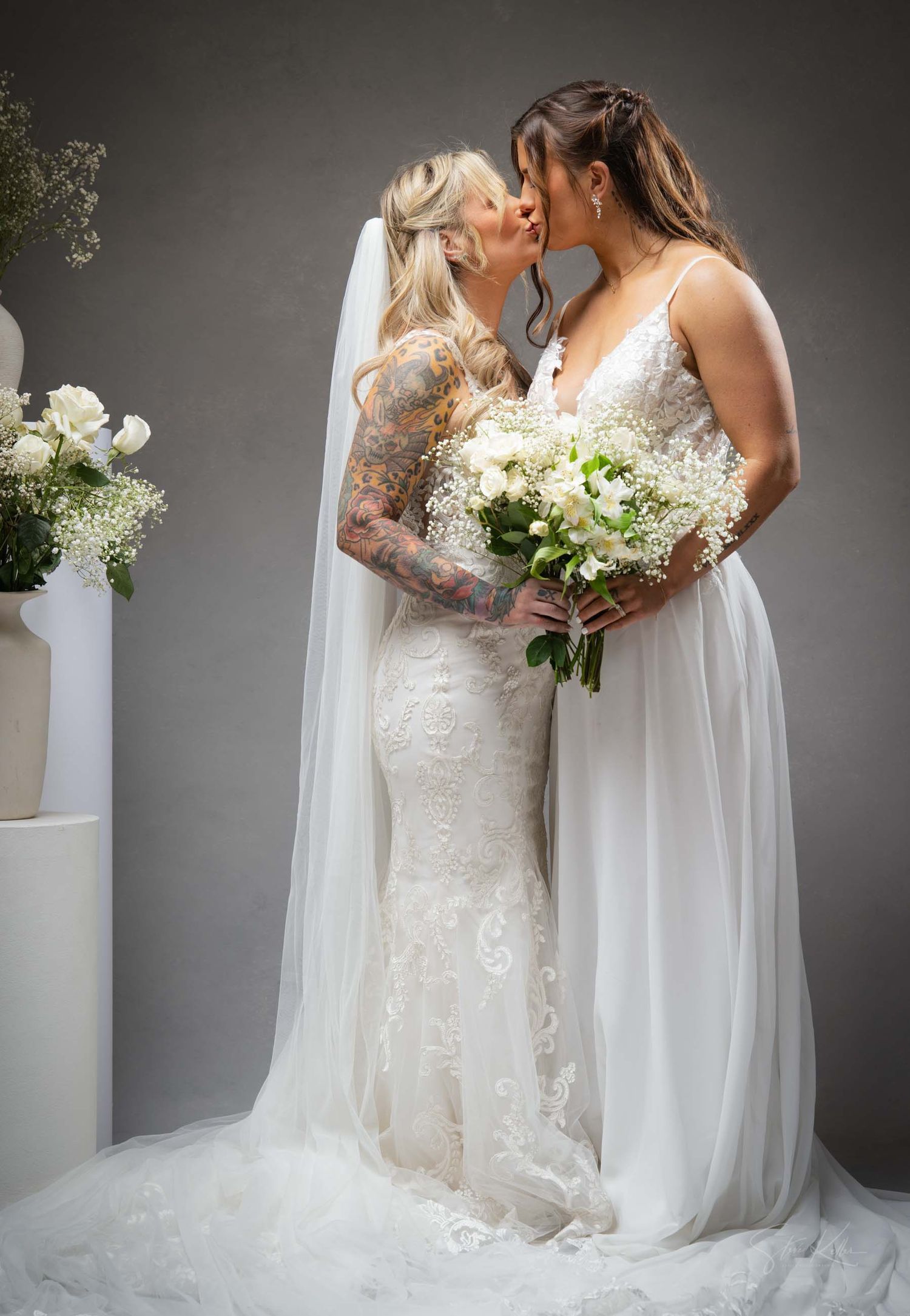 Two brides in elegant white wedding dresses share an intimate moment against a gray studio backdrop.