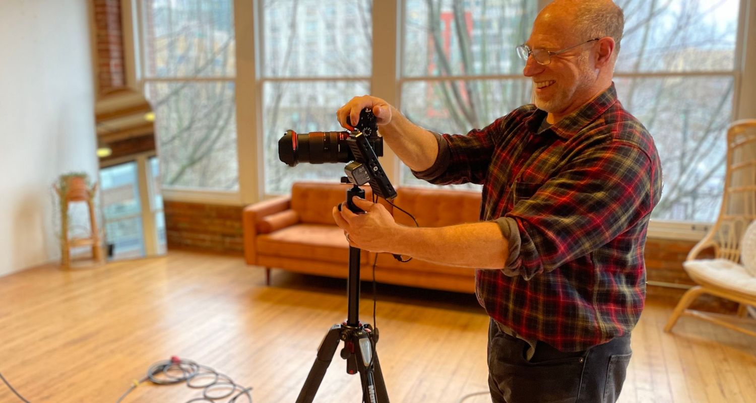 Smiling man in a plaid flannel shirt adjusts a camera mounted on a tripod in a bright, sunlit studio with hardwood floors.