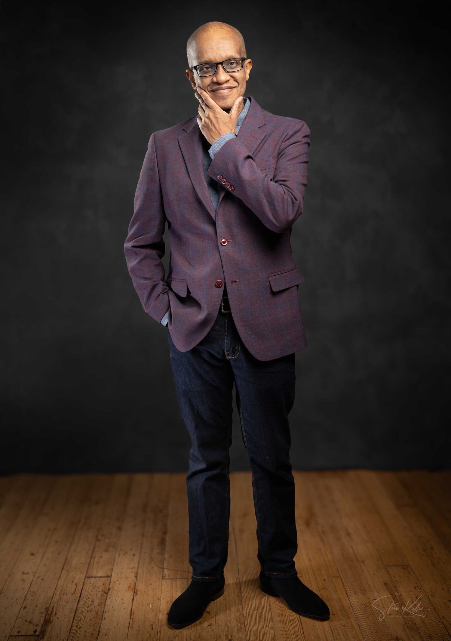 Bald man with glasses in purple blazer and jeans poses thoughtfully on hardwood floor against dark backdrop.