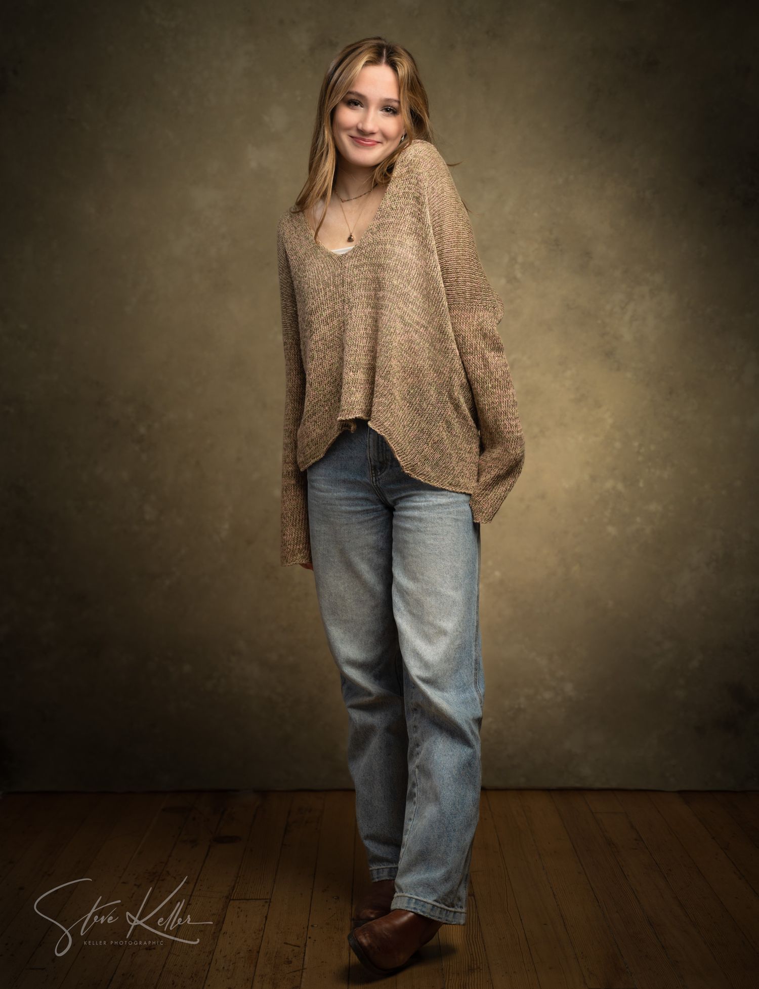 Young woman in beige knit sweater and light blue jeans poses for a professional studio portrait photo.