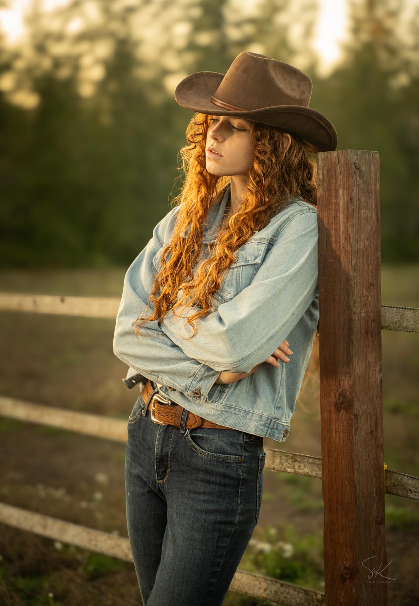 Red-haired woman in cowboy hat, denim jacket, and jeans leaning against wooden fence post at golden hour.