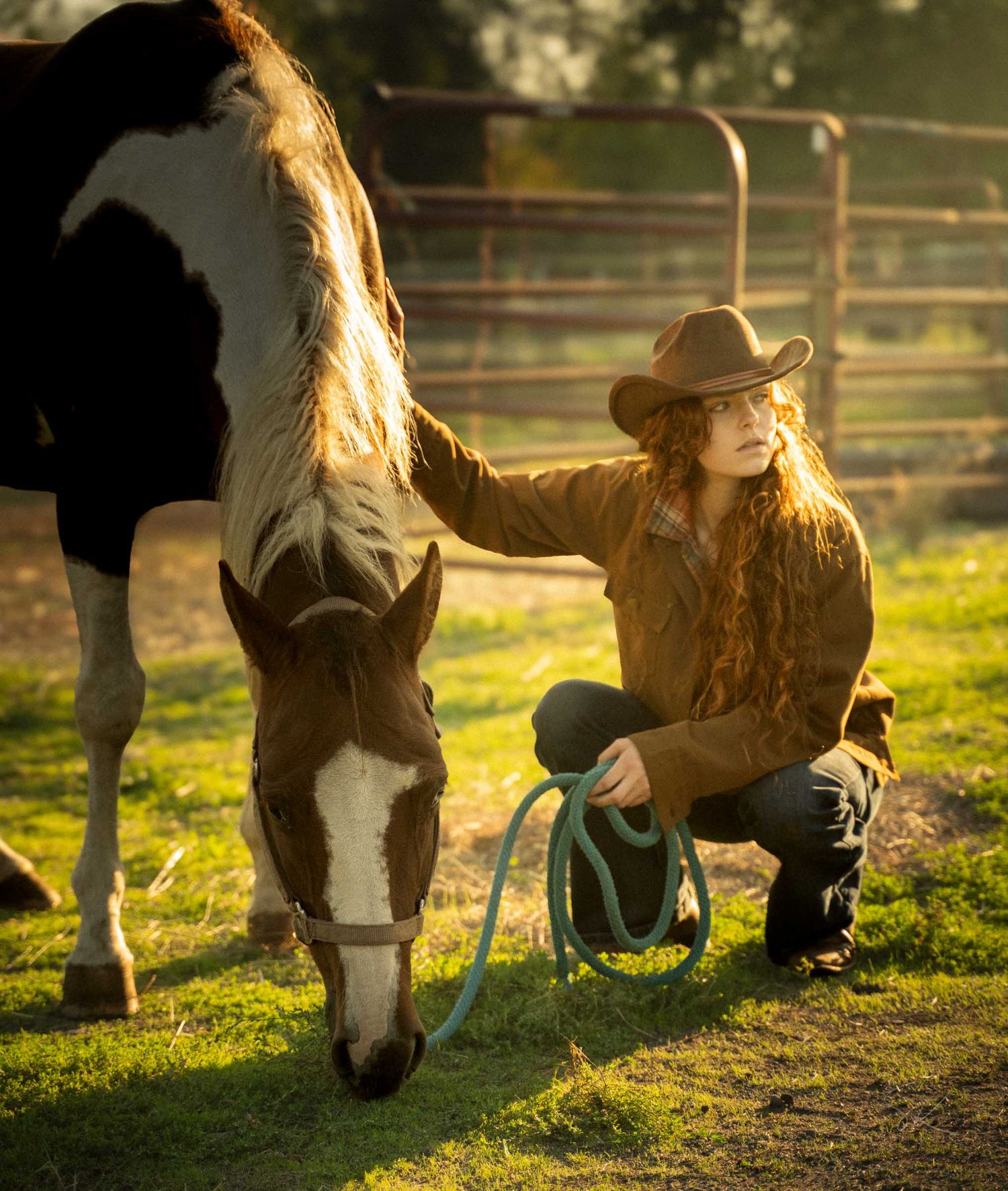 Woman in cowboy hat kneeling beside a paint horse on green grass at golden hour, holding a blue lead rope.